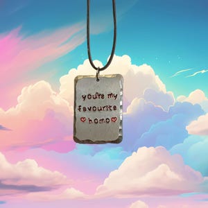 Peut inclure: Pendentif rectangulaire en argent avec l'inscription manuscrite "you're my favourite homo". Le mot "homo" est orné de deux cœurs rouges. Le pendentif est suspendu à un cordon noir sur fond de ciel.