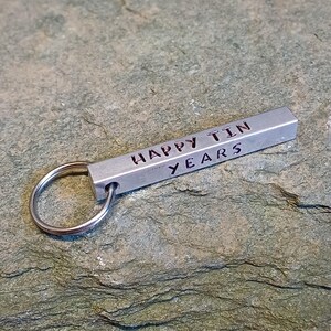 以下が含まれることがあります： 「Happy Tin Years」と書かれた長方形のバーが付いた銀色の金属製のキーホルダーです。