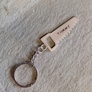 May include: Silver-colored keychain featuring a miniature saw design. The saw blade has a jagged edge and the word "TOMMY" is engraved on the handle. The keychain includes a chain and a circular key ring.