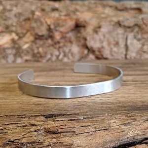 May include: A silver metal bracelet with a smooth, flat surface. The bracelet is open and has a simple design.