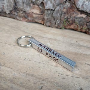 May include: A silver-toned metal bar keychain featuring a brushed finish and a circular split ring. The text "MARRIED AS FUCK" is stamped in dark, recessed lettering along the length of the rectangular bar, creating a clear and legible message.