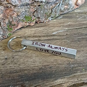 Puede incluir: Un llavero de metal plateado con una barra rectangular grabada con las palabras "IRON ALWAYS LOVE YOU" en letras rojas.