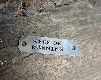 May include: A small, rectangular metal tag with the words "KEEP ON RUNNING" stamped in dark red letters. The tag has a hole on each end and is set against a wood background.