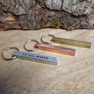 May include: Three metal keyrings in silver, copper, and brass. The silver keyring has the Roman numerals IV-VII-MMXX engraved on it. Each keyring has a rectangular bar attached to a silver ring.