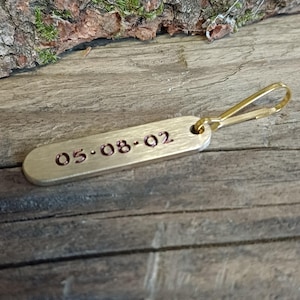 Pode incluir: Um chaveiro de prata com um fecho giratório dourado. A etiqueta é gravada com a data "05-08-02" em vermelho.