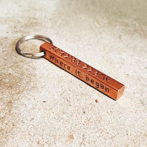 May include: A copper-coloured rectangular keyring with a silver key ring. The keyring is engraved with the text "52°42'33.223"W Where it began". The keyring is lying on a light grey surface.