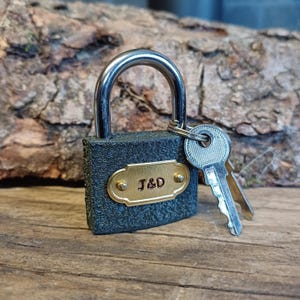 May include: A black padlock with a brass plate engraved with "J&D" and two keys. The padlock is sitting on a wooden surface.