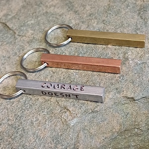 Könnte beinhalten: Drei Schlüsselanhänger aus Metall mit verschiedenen Oberflächen. Der obere Schlüsselanhänger ist golden, der mittlere ist kupferfarben und der untere ist silberfarben. Der silberne Schlüsselanhänger trägt die eingravierte Aufschrift "COURAGE DOESN'T".