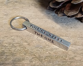 Op de afbeelding: Zilveren metalen sleutelhanger met een balk waarop "You're Steel The One" staat.