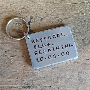 Könnte beinhalten: Ein rechteckiger Metall-Schlüsselanhänger mit einem silbernen Schlüsselring. Auf dem Schlüsselanhänger sind die Worte "REFERRAL. FLOW. REGAINING. 10.05.00" eingestanzt. Das Metall hat ein strukturiertes, gehämmertes Aussehen.