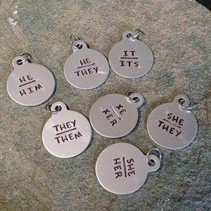 Może przedstawiać: Siedem srebrnych okrągłych znaczników z różnymi kombinacjami zaimków wygrawerowanych na nich. Znaczniki są wszystkie mniej więcej tego samego rozmiaru i mają otwór u góry do przymocowania do breloka lub innego przedmiotu. Tekst na znacznikach zawiera "On/On", "On/Oni", "Ona/Ona", "Ona/Oni", "Oni/Oni", "Ono/Ono" i "Xe/Xer".