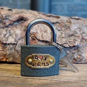 May include: A grey padlock with a silver shackle and a brass keyhole. The padlock has a brass plate with the inscription "D♥J Est '15" and a red heart.