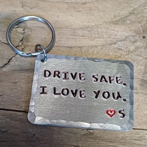 Può includere: Portachiavi in ​​argento con un'etichetta in metallo martellato che recita "Guida in sicurezza. Ti amo." con un piccolo cuore rosso e la lettera "S" sotto.