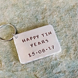 以下が含まれることがあります： 「Happy Tin Years 15.08.17」と刻まれた長方形のタグが付いたシルバーのキーホルダーです。