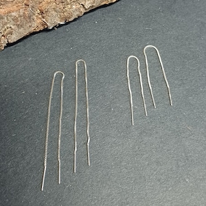 May include: A collection of silver threader earrings. The earrings are displayed on a dark gray surface, with a piece of wood in the background. The earrings have a delicate chain design, with a straight bar at the end.