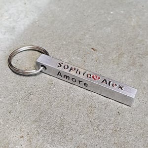 May include: Silver keychain with a rectangular bar engraved with the names "Sophie" and "Alex" separated by a red heart and the word "Amore".