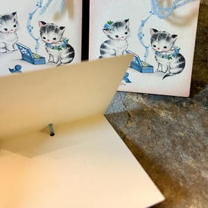 May include: Vintage greeting cards featuring illustrations of kittens. The kittens are depicted with blue floral accents and musical notes. Each card has a light blue string attached for hanging. The cards are on a pink background.