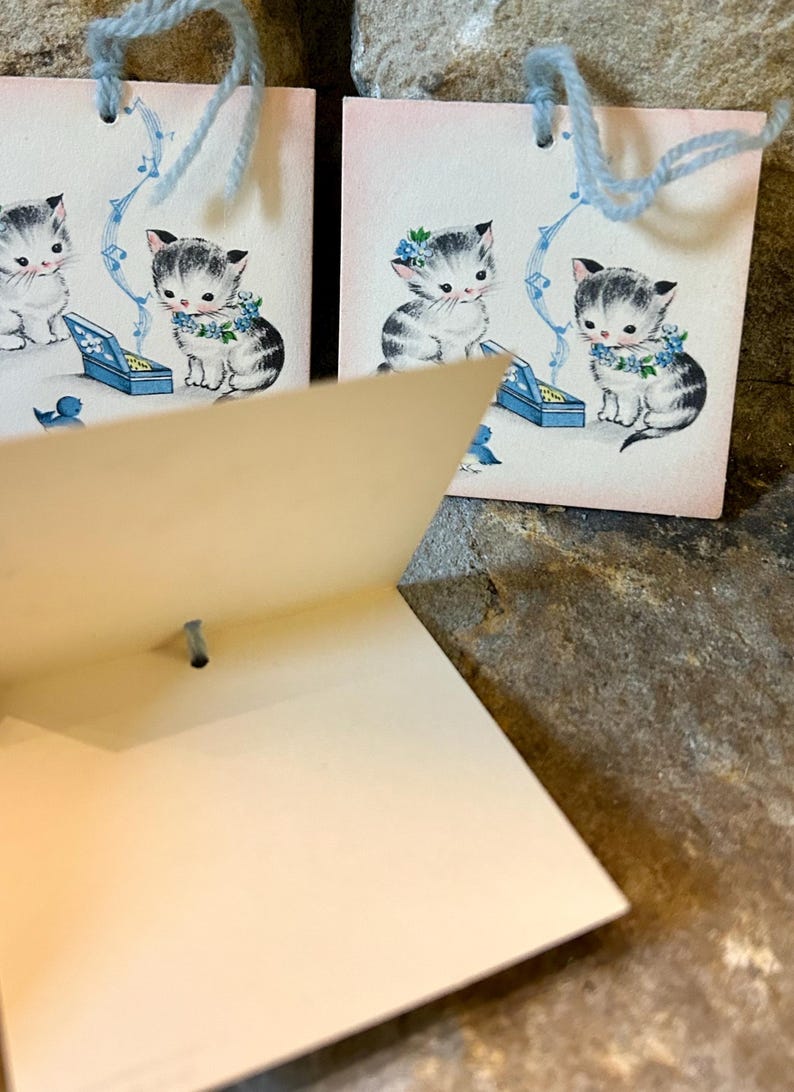 May include: Vintage greeting cards featuring illustrations of kittens. The kittens are depicted with blue floral accents and musical notes. Each card has a light blue string attached for hanging. The cards are on a pink background.