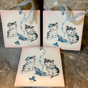 May include: Three square gift tags with a vintage illustration of kittens. Each tag features a kitten wearing a floral wreath, a blue bird, and a blue music box. The tags have light blue yarn handles and a pink background.