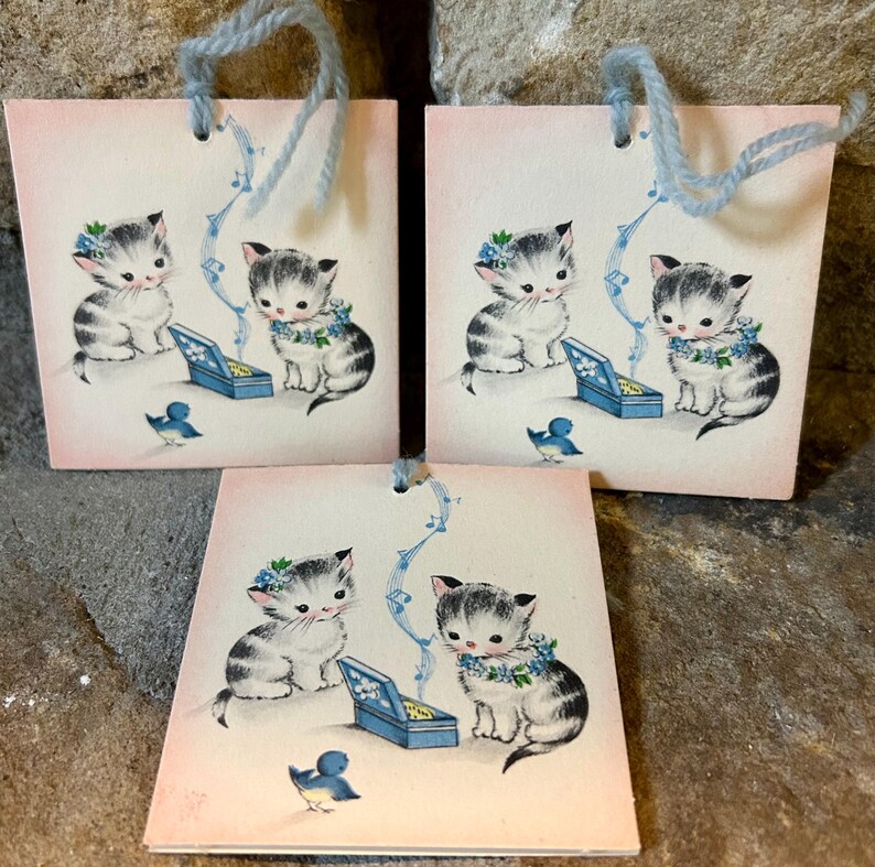 May include: Three square gift tags with a vintage illustration of kittens. Each tag features a kitten wearing a floral wreath, a blue bird, and a blue music box. The tags have light blue yarn handles and a pink background.