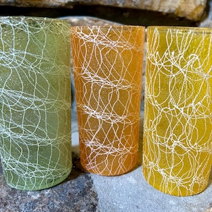 May include: Three cylindrical drinking glasses in shades of light green, orange, and yellow. Each glass features a textured, frosted surface overlaid with a white, chaotic, web-like pattern of thin, intersecting lines that wrap around the exterior.