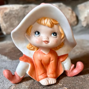 May include: A porcelain figurine of a girl elf wearing a white and pink hat, a red and orange dress, and white and pink shoes.