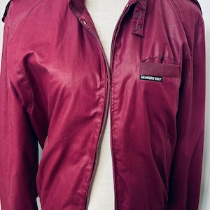 May include: A burgundy Members Only jacket featuring a high collar with a snap-button tab, a full-length front zipper, shoulder epaulets, and a small chest pocket with a black and white label that reads Members Only in white capital letters.