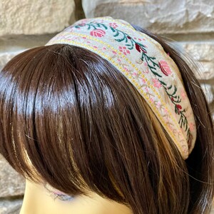 Op de afbeelding: Een beige hoofdband met een geborduurd bloemenmotief met roze, rode en groene bloemen. De hoofdband is gemaakt van een zachte, geweven stof.
