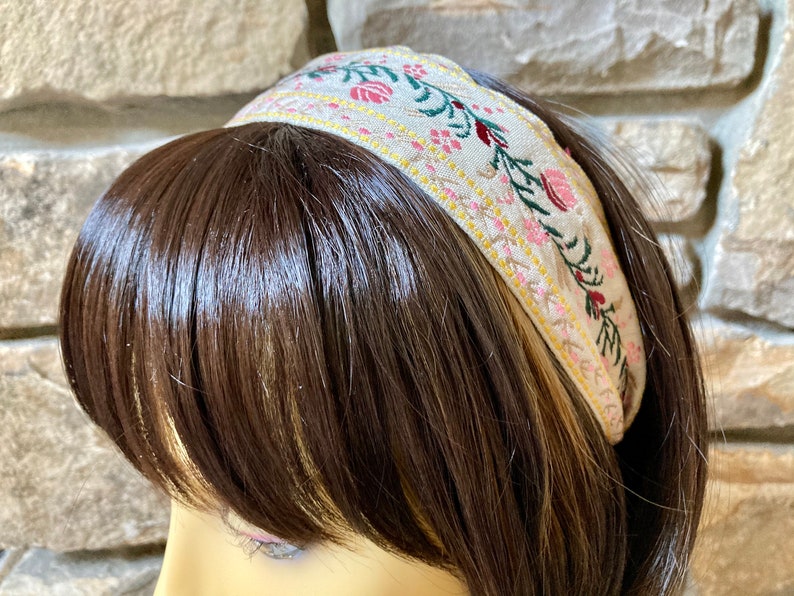 Op de afbeelding: Een beige hoofdband met een geborduurd bloemenmotief met roze, rode en groene bloemen. De hoofdband is gemaakt van een zachte, geweven stof.