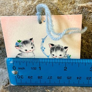 May include: A vintage gift tag featuring two adorable kittens with black and white fur, adorned with floral accents. The tag is approximately 2 inches wide and has a light blue yarn attached for hanging. The background is a soft pink.