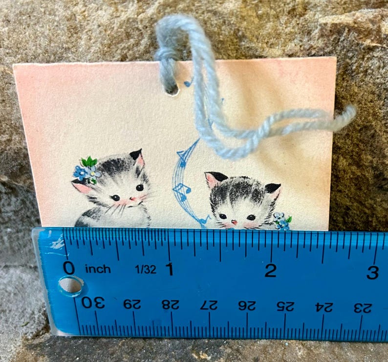 May include: A vintage gift tag featuring two adorable kittens with black and white fur, adorned with floral accents. The tag is approximately 2 inches wide and has a light blue yarn attached for hanging. The background is a soft pink.