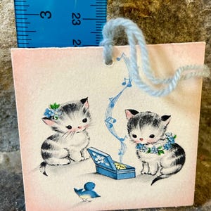 May include: A small, pink gift tag featuring two adorable kittens with black and white fur. The kittens are adorned with blue floral accents and are near a blue music box. A blue bird is also present. The tag has a light blue string.