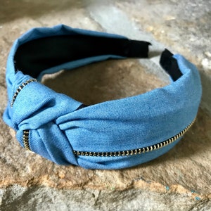 K&ouml;nnte beinhalten: Ein blaues Denim-Haarband mit einem goldenen Rei&szlig;verschlussdetail. Das Haarband ist oben gekn&uuml;pft und hat innen ein schwarzes Gummiband.