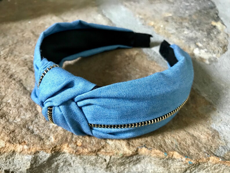 K&ouml;nnte beinhalten: Ein blaues Denim-Haarband mit einem goldenen Rei&szlig;verschlussdetail. Das Haarband ist oben gekn&uuml;pft und hat innen ein schwarzes Gummiband.