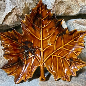 May include: A ceramic leaf-shaped dish with a glossy, brown finish. The dish has a detailed vein pattern and a stem. The edges of the leaf are jagged. The dish is likely used for serving or decoration.