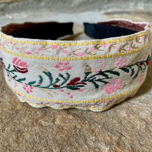 Op de afbeelding: Een beige hoofdband met een bloemenpatroon in roze, groen en bordeauxrood. De hoofdband heeft een gele rand.