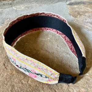 Op de afbeelding: Een beige hoofdband met een bloemenpatroon in roze, geel en groen. De hoofdband heeft een zwart elastiek aan de achterkant.