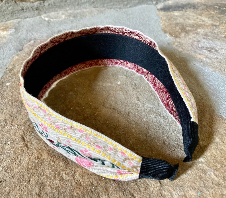 Op de afbeelding: Een beige hoofdband met een bloemenpatroon in roze, geel en groen. De hoofdband heeft een zwart elastiek aan de achterkant.