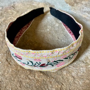 Op de afbeelding: Een beige hoofdband met een bloemenpatroon in roze, geel en groen. De hoofdband is gemaakt van stof en heeft een zwarte elastische band aan de binnenkant.