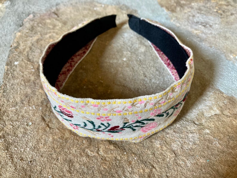 Op de afbeelding: Een beige hoofdband met een bloemenpatroon in roze, geel en groen. De hoofdband is gemaakt van stof en heeft een zwarte elastische band aan de binnenkant.