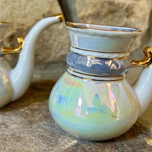 May include: Two white ceramic pipe-shaped decorative pieces with iridescent finishes. Each has a bulbous base, a light blue band, and gold accents on the handle and rim. The curved stem extends from the top.