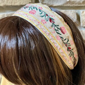 Op de afbeelding: Een beige hoofdband met een bloemenpatroon in roze, groen en rood. De hoofdband is gemaakt van stof en heeft een subtiele glans.
