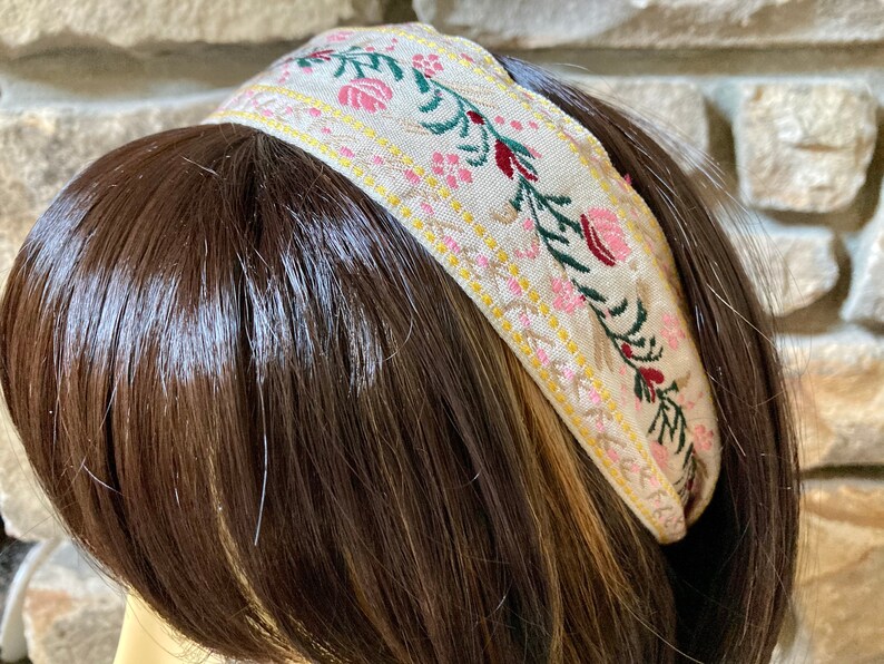 Op de afbeelding: Een beige hoofdband met een bloemenpatroon in roze, groen en rood. De hoofdband is gemaakt van stof en heeft een subtiele glans.