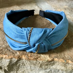 K&ouml;nnte beinhalten: Ein hellblaues Stirnband mit einem goldenen Rei&szlig;verschlussdetail. Das Stirnband ist in der Mitte geknotet und hat ein schwarzes Gummiband auf der R&uuml;ckseite.