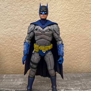 Puede incluir: Una figura de acción de Batman en azul y gris, con una capa azul y un cinturón de herramientas amarillo. La figura tiene una escultura detallada y está en posición de pie.
