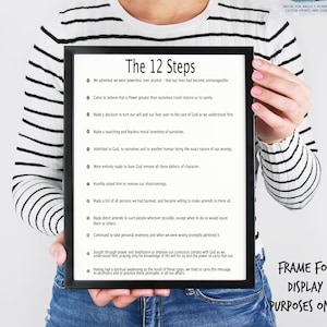 May include: A framed print with the text "The 12 Steps" in a black frame. The text lists the twelve steps of recovery, with each step numbered and beginning with a bullet point. The print is held by a person wearing a striped shirt.