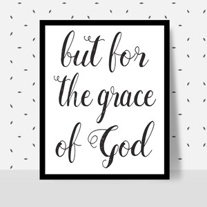 May include: A framed print featuring the phrase "but for the grace of God" in black cursive lettering. The text is set against a white backdrop, enclosed within a black frame. The background has a pattern of small grey leaf shapes.
