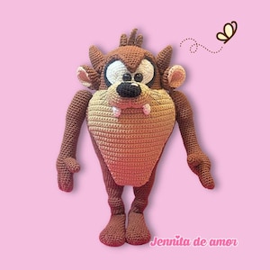 Op de afbeelding: Een bruin en beige gehaakt knuffeldier dat lijkt op het personage de Tasmaanse Duivel. Het heeft grote witte ogen met zwarte pupillen, een zwarte neus, twee kleine roze hoektanden en puntige oren. De tekst Jennita de amor is in het roze gedrukt.