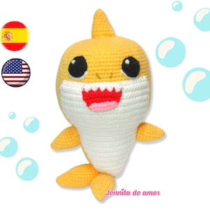 Puede incluir: Un juguete de tiburón amarillo de crochet con dientes blancos y grandes ojos negros. El tiburón tiene la barriga blanca y sostiene un corazón rosa en la boca. El juguete está rodeado de burbujas azules.