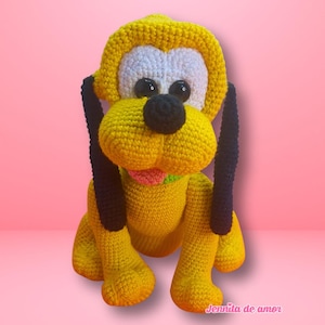 Puede incluir: Un juguete de perro hecho a mano con ganchillo, en posición sentada. El perro es principalmente amarillo con orejas negras y una nariz negra. Los ojos son negros y la boca rosa. El juguete tiene una cara blanca y está sobre un fondo rosa.